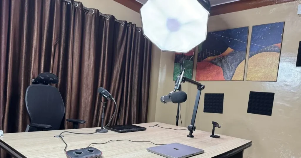 Estúdio de podcast com dois microfones, iluminação de teto, painel acústico e mesa com notebook.


