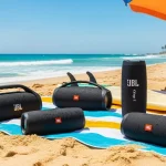 Cinco caixas de som JBL dispostas sobre canga listrada na praia com mar azul turquesa ao fundo