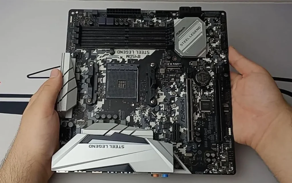 Pessoa segurando a placa-mãe ASRock B450M Steel Legend, modelo Micro-ATX com design camuflado em preto, branco e cinza.

